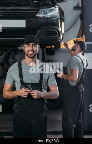 Mécanicien automobile dans des combinaisons clés holding, tandis que collègue travaillant en atelier derrière Banque D'Images