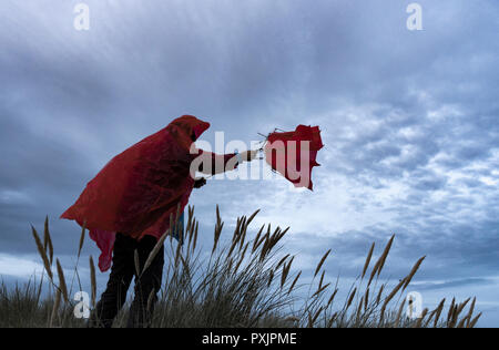 Homme portant un manteau de pluie imperméable rouge luttant avec un parapluie par temps venteux. Banque D'Images
