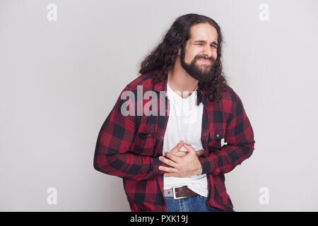 Portrait d'un bel homme avec barbe et cheveux longs noirs dans un style décontracté, chemise rouge à carreaux debout tenant son ventre avec douleur. Piscine Banque D'Images