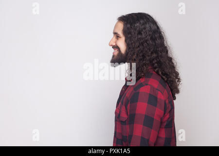 Vue côté profil portrait of handsome homme heureux avec barbe et black long cheveux bouclés dans un style décontracté, chemise rouge à carreaux avec permanent sourire à pleines dents. Banque D'Images