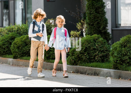 Cute little schoolkids avec sacs à dos se tenant la main et marcher ensemble sur la rue Banque D'Images