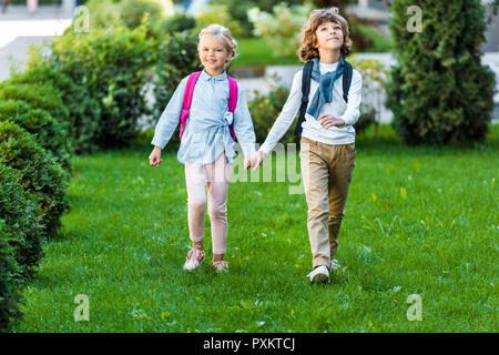 Sur toute la longueur de l'adorable heureux schoolkids with Backpacks holding hands sur pelouse verte Banque D'Images