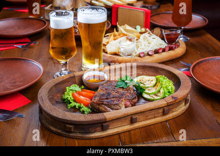 Sex steak avec tomates, salade, courgette, verts et de la sauce sur une planche de bois. Deux verres de bière brune Banque D'Images