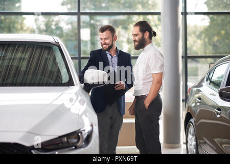 Manager du dossier et montrant pour le client d'une voiture automobile blanc centre. Hommes debout près de l'auto, l'examinant et en souriant. Voiture de porter en veste bleu foncé, l'acheteur en chemise blanche. Banque D'Images