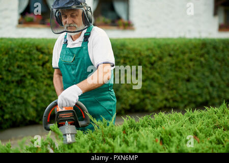 Senior male gardener clipping en couverture, le jardin à l'aide du coupe-haie d'essence. Jardin professionnel worker wearing en uniforme vert avec un casque de protection et masque de sécurité travaillant avec massicot. Banque D'Images