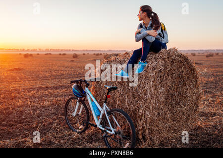 Les jeunes cyclistes d'avoir se reposer après une promenade en terrain d'automne au coucher du soleil. Woman admiring view sitting on haystack. Sport Loisirs concept Banque D'Images