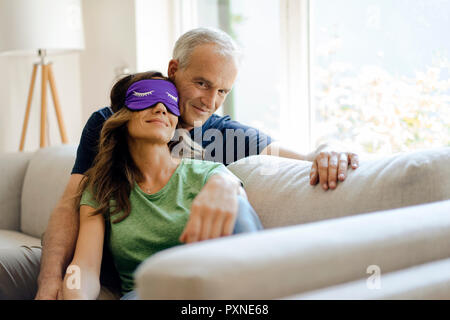 Smiling young couple sitting on couch à la maison avec femme portant un masque pour les yeux Banque D'Images