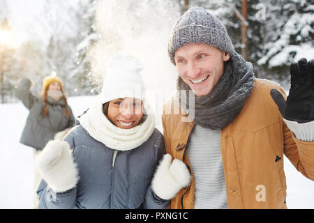 Cheerful heureux jeune multi-ethnic couple dans des foulards et des chapeaux de la protection de la neige jeté par des amis lors de la marche en forêt Banque D'Images