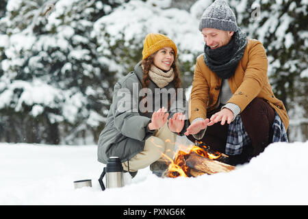 Cheerful beau jeune couple assis par puffy jackets fire et se réchauffer les mains tout en profitant de la forêt en hiver date romantique Banque D'Images