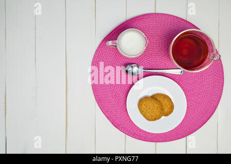 Du thé chaud, des biscuits et du lait sont disposés sur un napperon rose sur un tableau blanc, tableau. L'arrangement est décentré. Banque D'Images