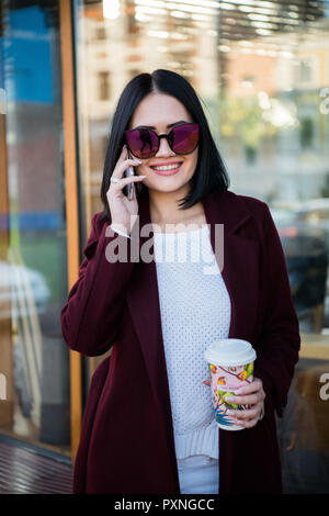 La mode Smiling Woman talking on mobile phone et de boire du café à emporter dans la ville. Banque D'Images