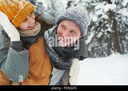 Smiling beau jeune homme en bonnet et écharpe en tricot copine donnant piggyback ride en forêt d'hiver, elle réglage hat Banque D'Images