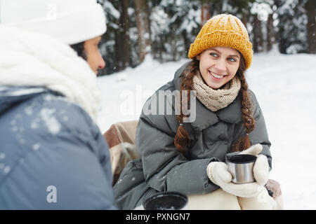 Jolie fille positive en tricot jaune hat parler à tes amis et boire le thé de thermos mug au camping en forêt d'hiver Banque D'Images