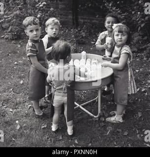 Années 1950, les jeunes enfants à une crèche ou à l'extérieur de l'école maternelle de s'amuser ensemble jouant avec de vieilles bouteilles de lave et l'eau dans un grand bol, une activité qui favorise la découverte sensorielle et du développement des compétences. Banque D'Images