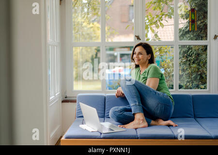 Smiling mature woman sitting on sofa at home with laptop Banque D'Images