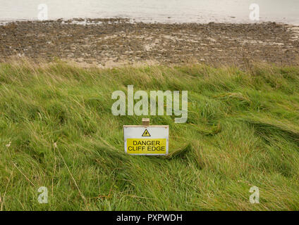 Panneau d'avertissement jaune et blanc de bord de falaise danger sur le chemin de la côte du pays de galles, plage, marée basse en arrière-plan, érosion côtière près de Pwllheli côte nord du pays de galles uk Banque D'Images