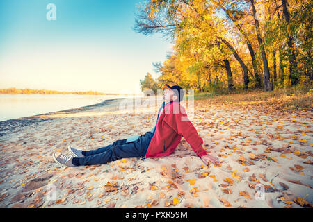 Un homme heureux avec les yeux fermé assis sur la rive du fleuve sur le sable avec des feuilles tombées en automne. L'homme en bonne météo ensoleillée Banque D'Images