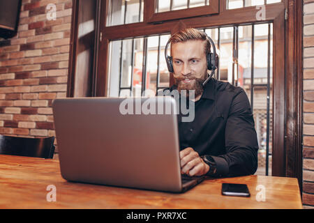 Homme barbu souriant avec Travail avec casque avec laptop in cafe Banque D'Images