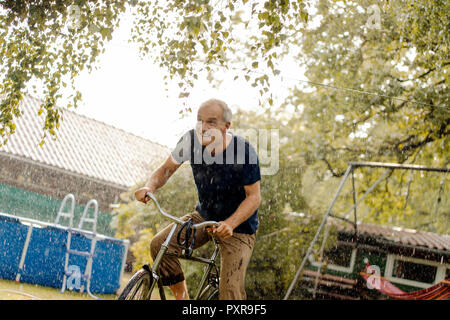 Happy man riding bicycle in summer rain in garden Banque D'Images