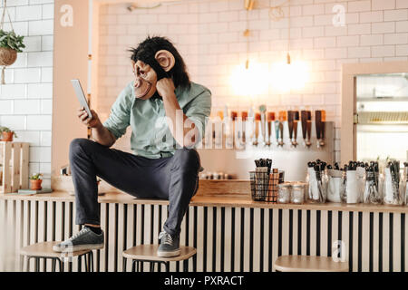 Homme portant un masque singe, assis sur le comptoir d'un bar, using digital tablet Banque D'Images