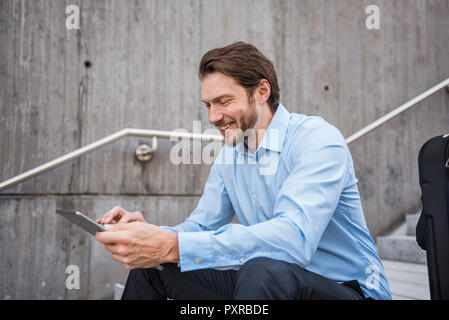 Smiling businessman with suitcase sitting on stairs using tablet Banque D'Images