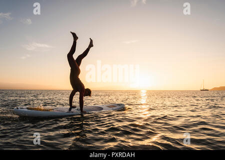 Young man doing handstand sur paddleboard au coucher du soleil Banque D'Images