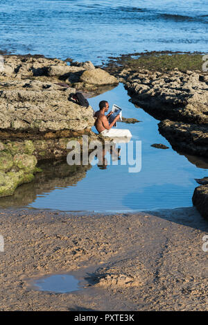Estoril, Portugal - Juillet 9, 2018 : l'homme en pantalon de lin assis dans waterfilled rock couverte par la mer de l'Atlantique la lecture d'un journal dans l'après-midi sunsh Banque D'Images