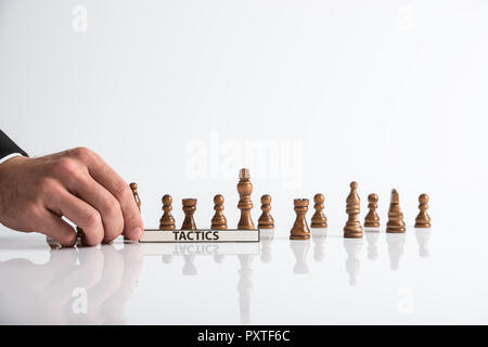 Chef d'entreprise titulaire d'un signe de tactiques en face de pièces d'échecs en bois placées sur blanc 24 avec réflexion. Banque D'Images