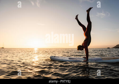 Young man doing handstand sur paddleboard au coucher du soleil Banque D'Images