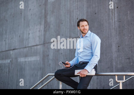 Businessman with smartphone et écouteurs assis sur une balustrade Banque D'Images