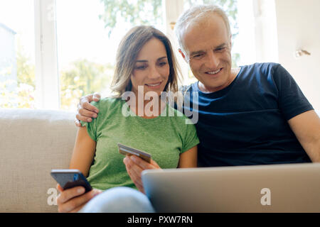 Smiling young couple sitting on sofa at home achats en ligne avec laptopand téléphone intelligent Banque D'Images