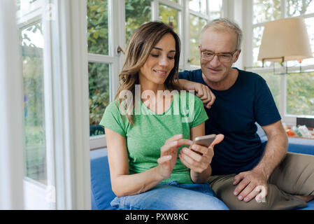 Smiling young couple sitting on couch au partage à domicile cell phone Banque D'Images