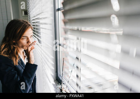 Femme debout par fenêtre, espionnage through blinds Banque D'Images