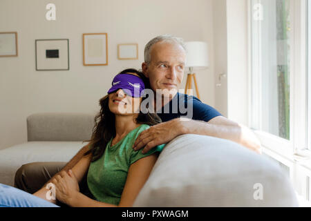 Smiling young couple sitting on couch à la maison avec femme portant un masque pour les yeux Banque D'Images