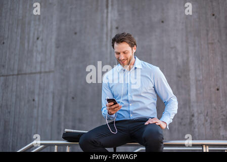 Smiling businessman with earbuds assis sur une balustrade looking at smartphone Banque D'Images