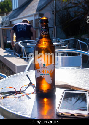 Bouteille de bière, Cornish Korev golden rougeoyant dans la lumière du soleil sur une table à l'extérieur avec des lunettes et un iPhone comme leur propriétaire se détend dans la chaleur. Banque D'Images