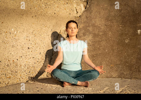 Femme pratiquant le yoga et la méditation dans le soleil par un mur de béton Banque D'Images