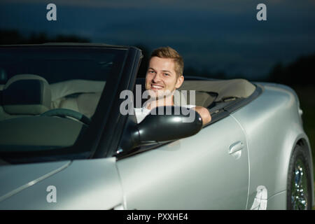 Bel homme assis sur le siège du conducteur, de l'argent cabriolet et posant. Conducteur looking at camera et souriant. Concept d'automobiles de luxe et les voyages en voiture. Banque D'Images