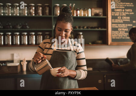 Smiling young African barista de verser du lait dans un cappuccino Banque D'Images