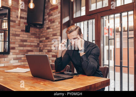 Jeune homme triste barbu est assis à table, couvrant son visage avec sa main et on cell phone. Le bureau est l'ordinateur portable, tablettes. Man Banque D'Images