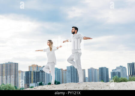 Belle jeune couple practicing yoga Yoga en posture de l'arbre (Vrksasana) sur sandy dune avec des bâtiments de la ville en arrière-plan Banque D'Images