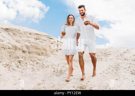 Happy young couple en blanc avec des cocktails de coco marche sur les dunes de sable Banque D'Images