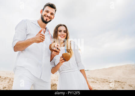 Happy young couple en blanc avec des cocktails de coco looking at camera Banque D'Images