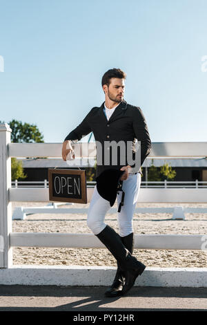 Beau mâle equestrian leaning on fence et holding open sign at horse club Banque D'Images