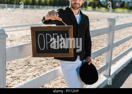 Portrait d'homme equestrian leaning on fence et montrant open sign at horse club Banque D'Images