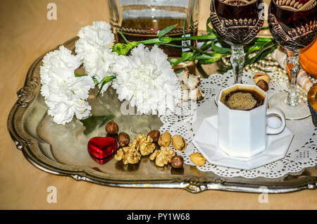 Un plateau d'argent nickel avec un bouquet d'œillets, de café noir,vieux verres en cristal et une bouteille de liqueur, chocolat coeur,écrous et une poire,orange Banque D'Images