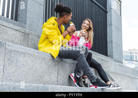 Smiling multicultural couple eating hamburgers près de skateboard au city street Banque D'Images