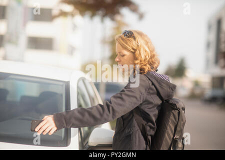 Femme debout devant une voiture en ouvrant la porte avec son téléphone portable Banque D'Images