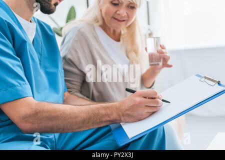 Cropped shot of male nurse writing on clipboard et senior woman drinking water Banque D'Images