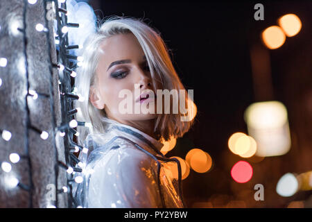 Beau young woman leaning on wall with white garland on city street at night Banque D'Images